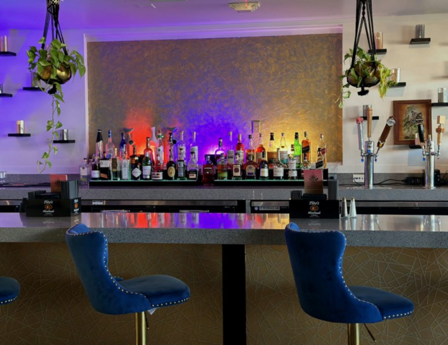 The bar at Saffron, with bottles backlit against a gold-textured wall under purple light, blue velvet stools along the stone counter, and hanging plants at either side