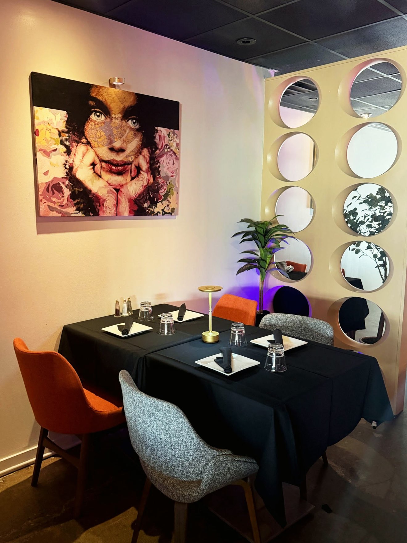 Corner table for four set with black linen under a floral portrait canvas, beside a wall of circular mirrors and a warm brass table lamp