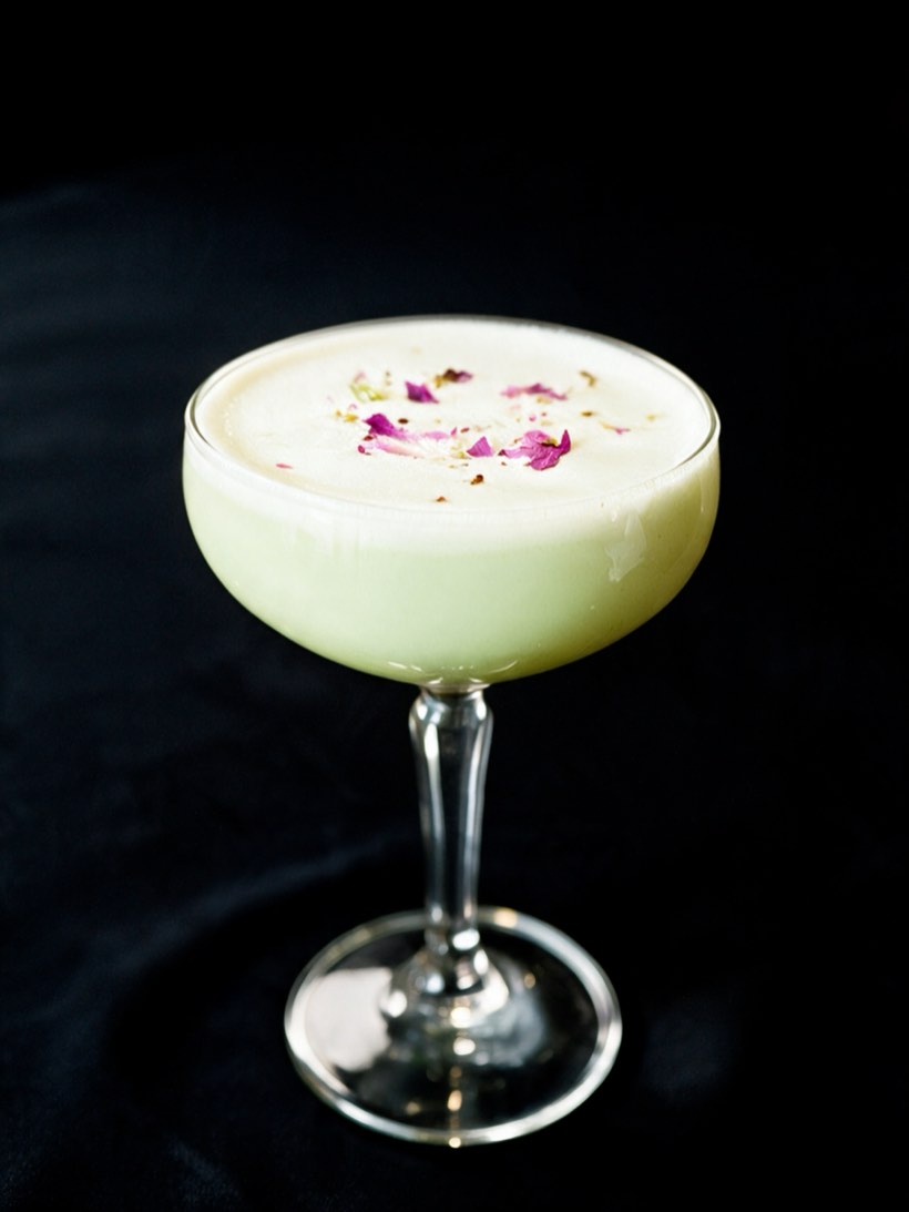 Pale green Naughty Coconut in a stemmed coupe under a thick foam cap scattered with rose petals and crushed pistachio