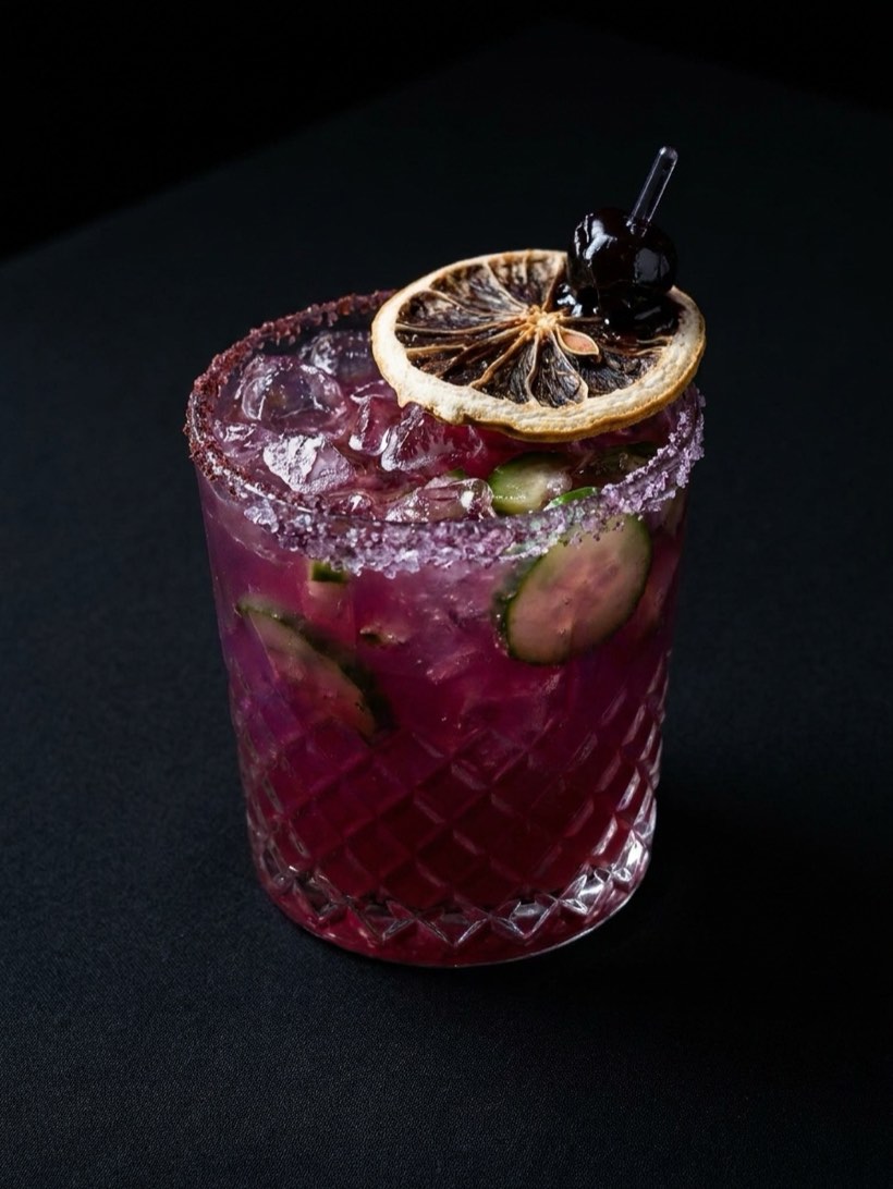 Magenta pomegranate margarita over crushed ice with cucumber slices, a sugared rim, dried citrus and a dark cherry