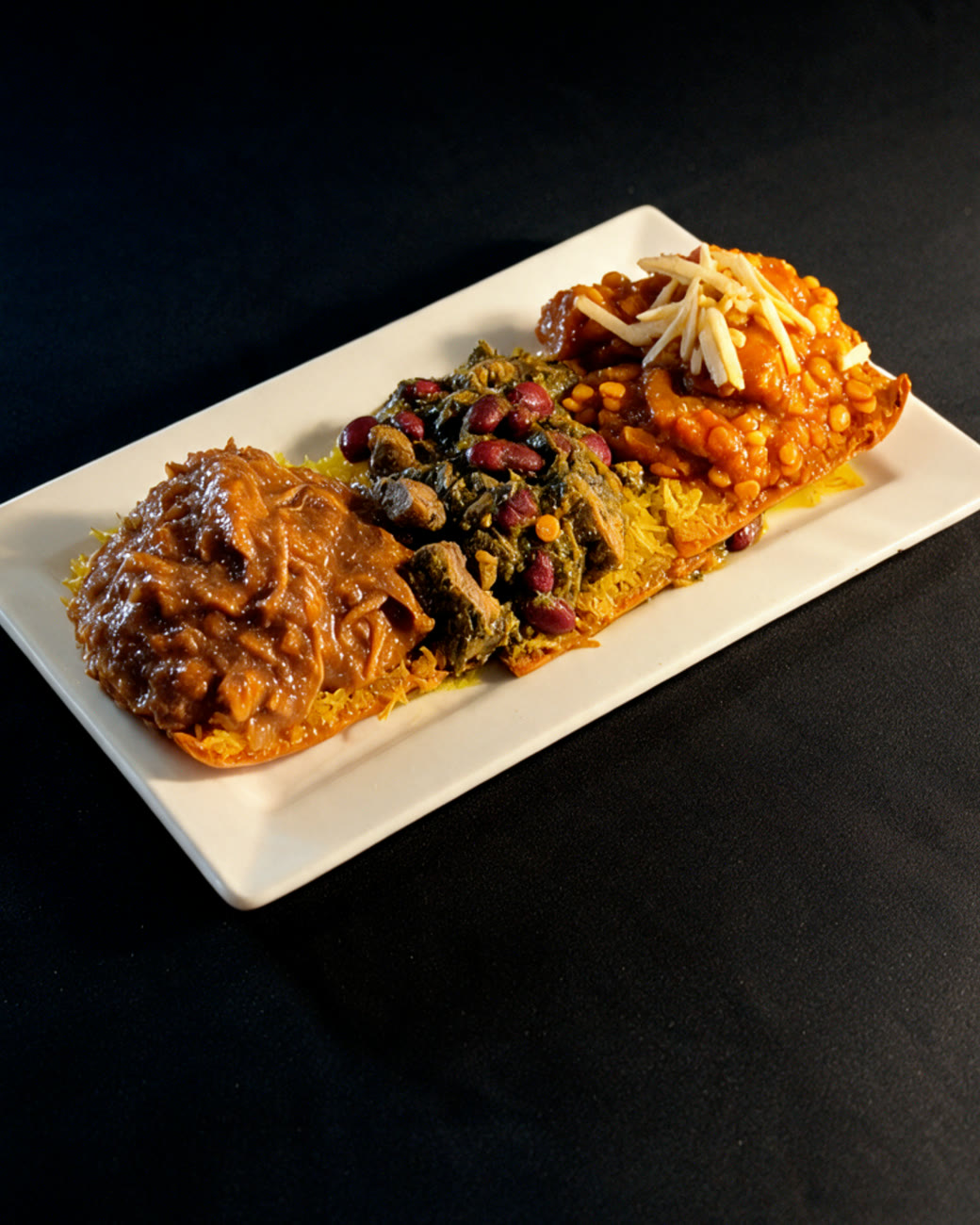 A long white platter of crispy tahdig topped with three house stews: fesenjoon, ghormeh sabzi and gheymeh bademjan